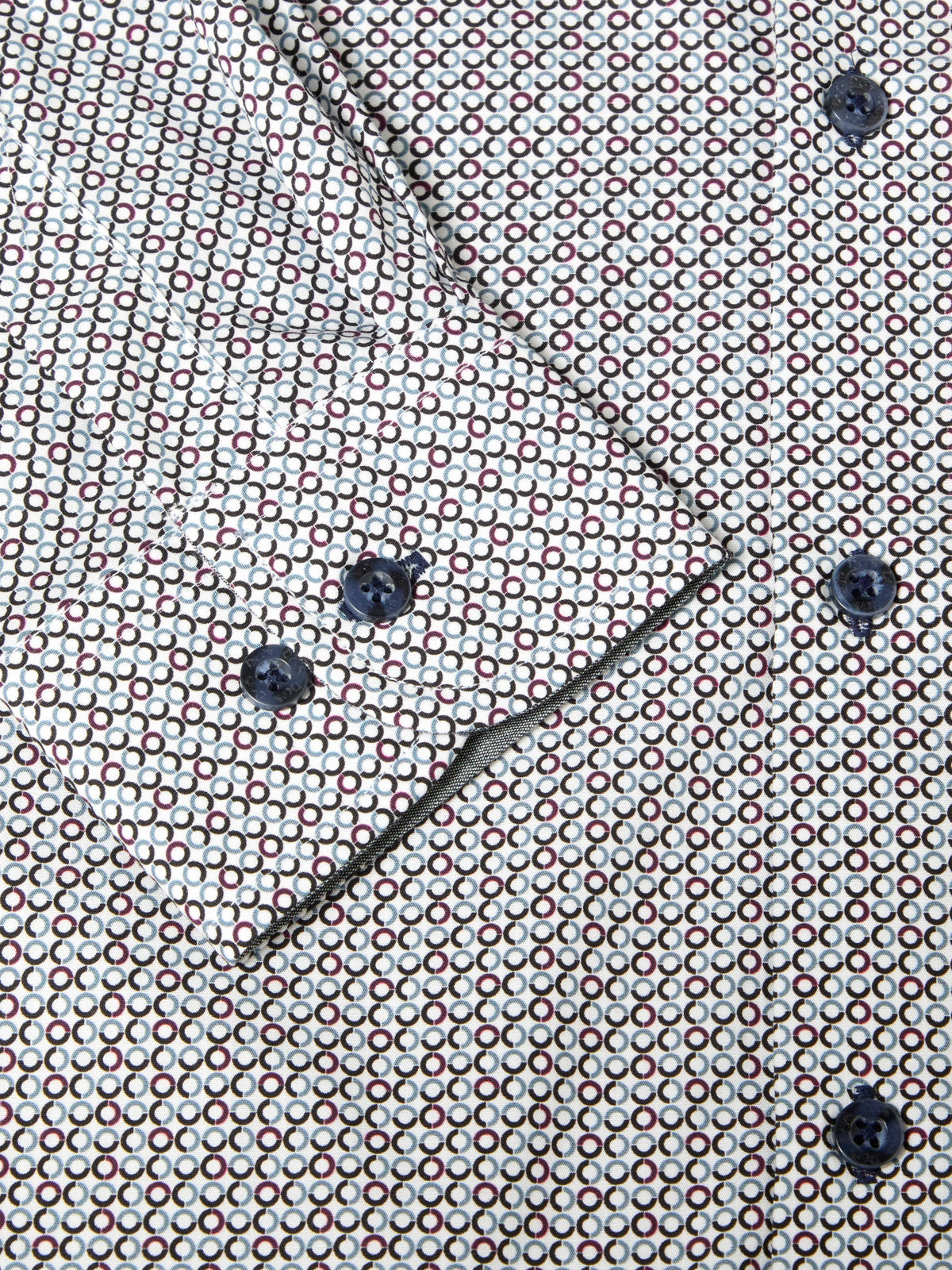 Close-up of a patterned shirt fabric with a repeating small geometric motif in red, white, and dark blue, plus matching dark buttons and a diagonal fold.