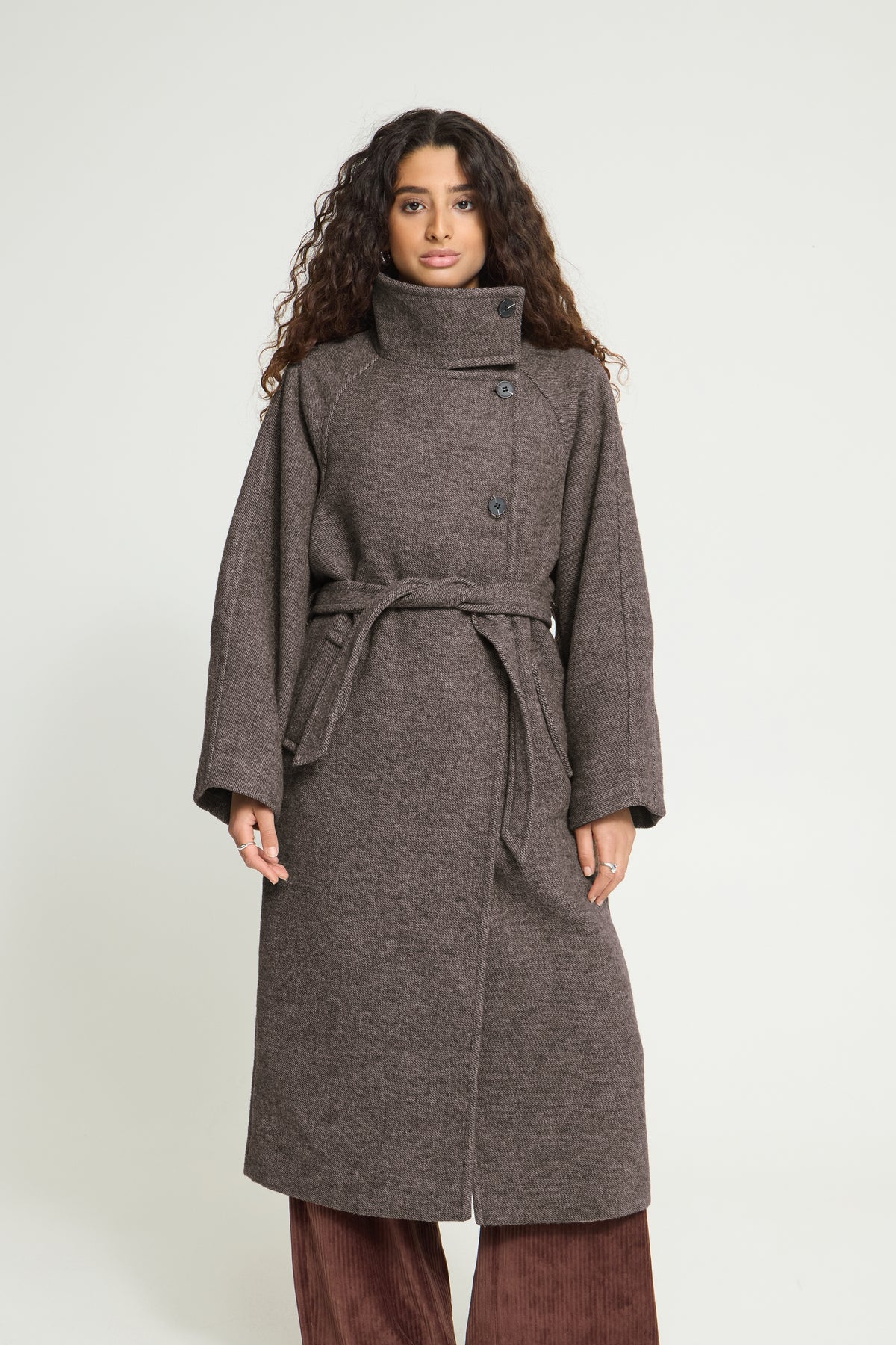 A woman modeling a mid-length, dark brown textured coat with a high collar, button front, and matching waist tie, paired with brown pants against a light gray studio background.