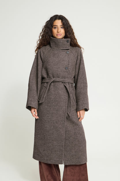 A woman modeling a mid-length, dark brown textured coat with a high collar, button front, and matching waist tie, paired with brown pants against a light gray studio background.