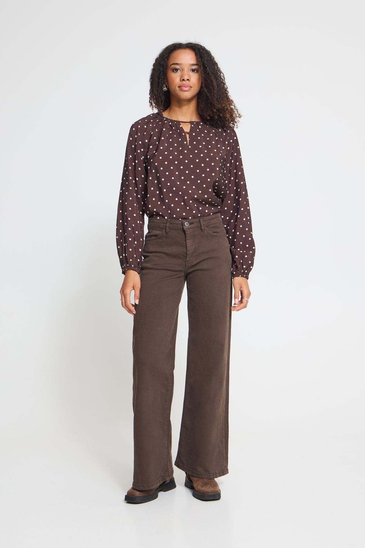A woman stands against a light gray studio background wearing a dark brown long-sleeve blouse with small white polka dots and high-waisted, wide-leg brown pants.