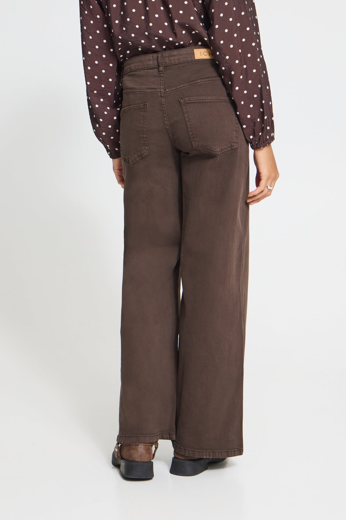 A person shown from the back wearing dark brown wide-leg jeans, a brown polka-dot blouse, and dark shoes against a plain light gray studio background.
