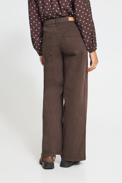 A person shown from the back wearing dark brown wide-leg jeans, a brown polka-dot blouse, and dark shoes against a plain light gray studio background.