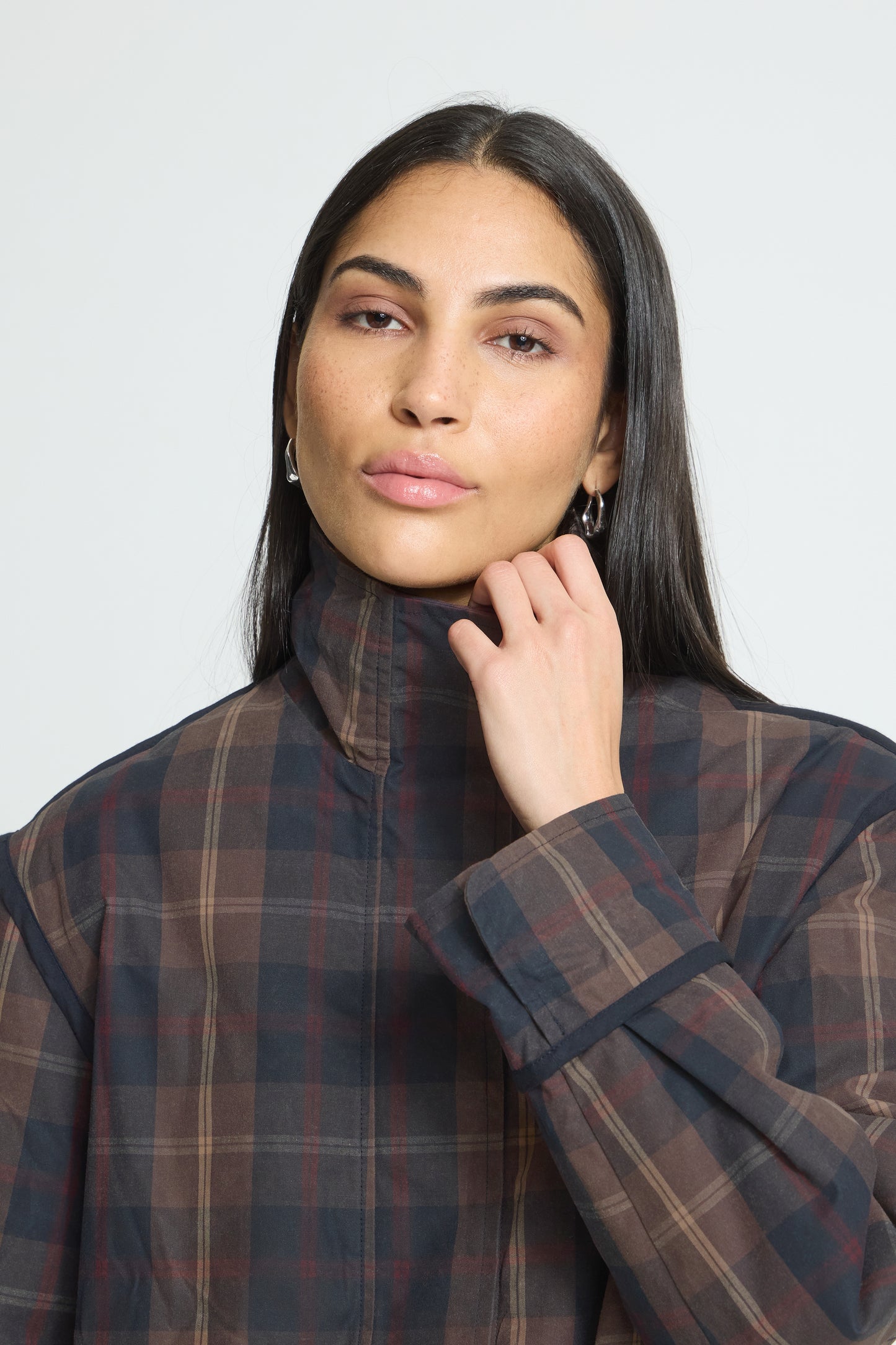 A woman with long dark hair wearing a dark brown and black plaid high-neck jacket, posing against a light gray studio background.