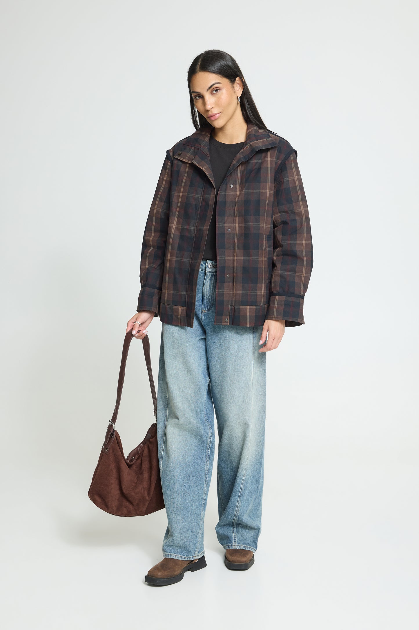 A woman stands against a light gray studio background wearing a dark brown and black plaid hooded jacket over a black top, light blue wide-leg jeans, and brown shoes, while holding a brown shoulder bag.