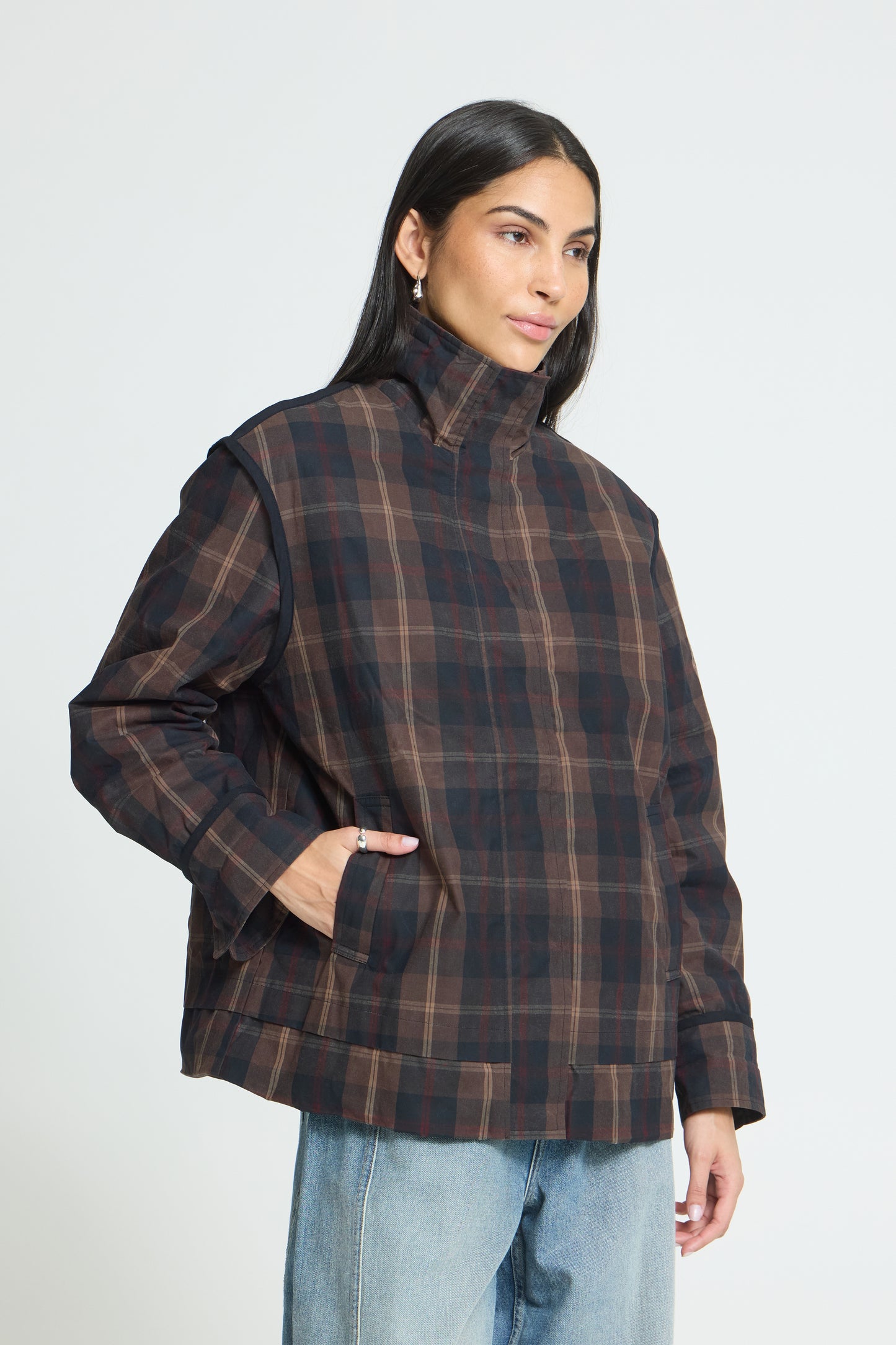 A woman wearing a dark brown and black plaid jacket with a high mock neck, paired with light blue jeans, photographed against a light gray studio background.