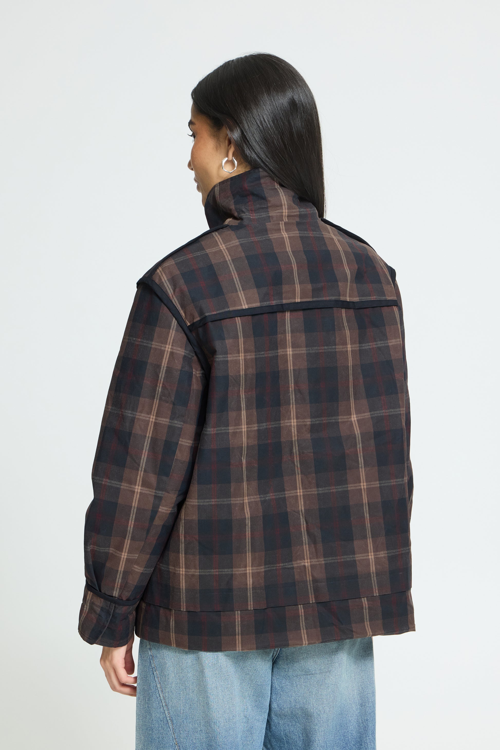 A woman shown from the back wearing a dark brown and black plaid jacket with a high collar, paired with light blue jeans, against a plain light gray background.
