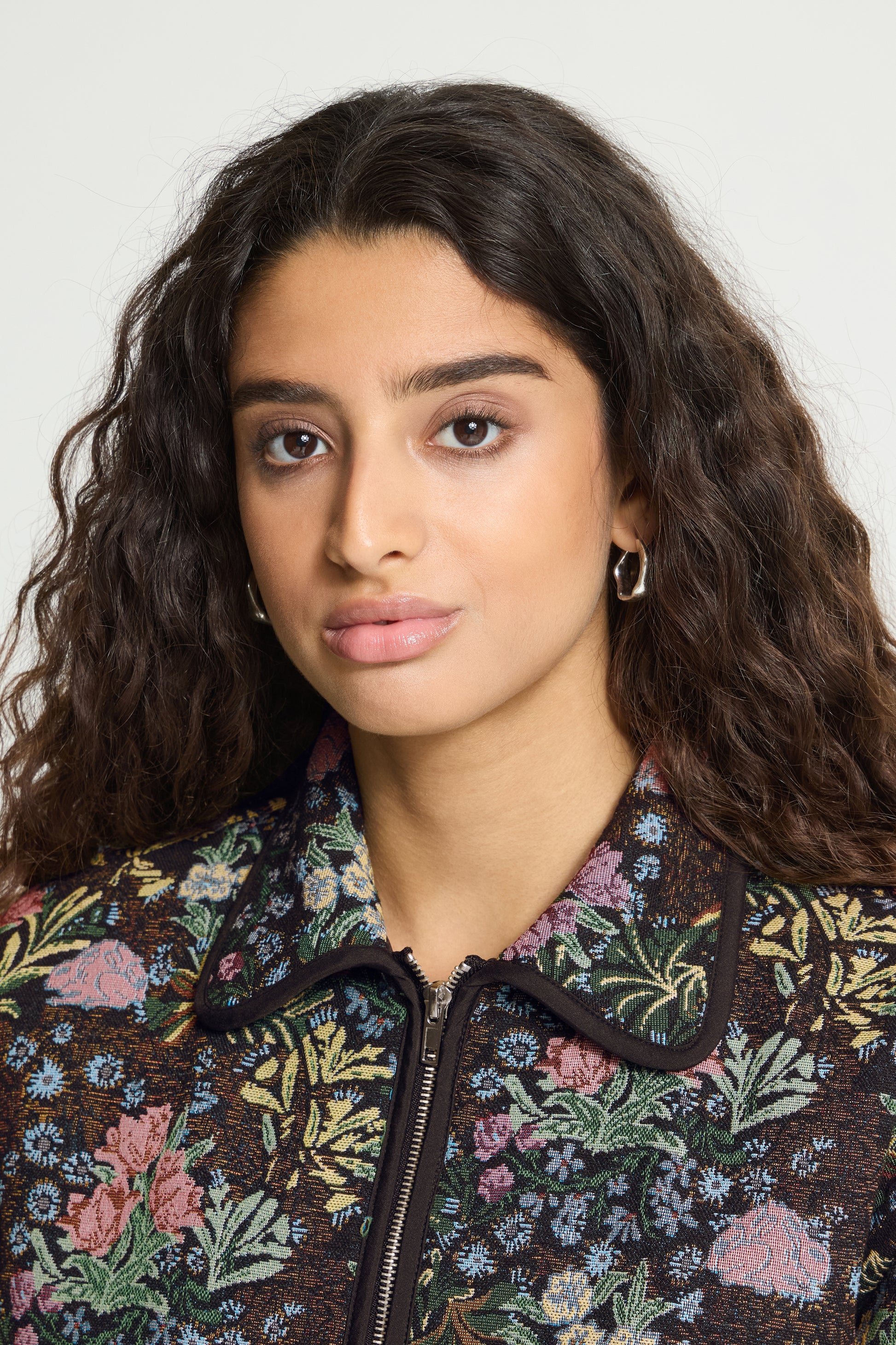 A close-up portrait of a woman with dark curly hair wearing a dark floral embroidered jacket with a collar and zipper, set against a plain light gray background.