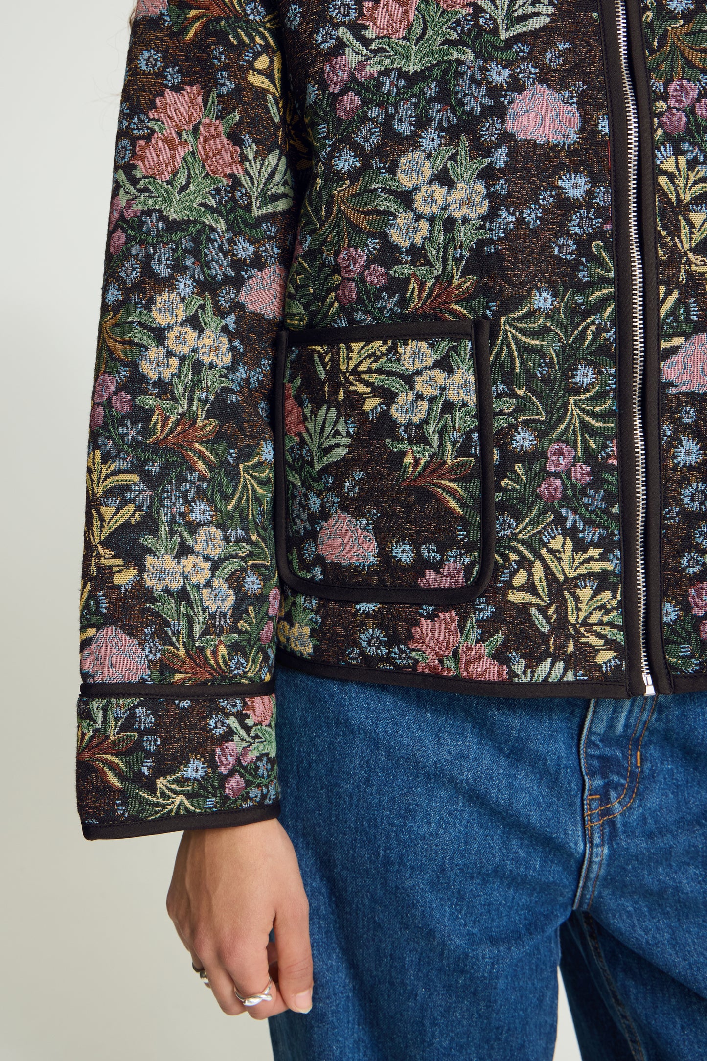 A close-up of a person wearing a dark floral quilted jacket with pink, green, and blue flowers, paired with blue jeans.