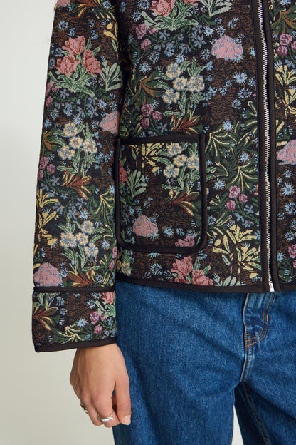 A close-up of a person wearing a dark floral quilted jacket with pink, green, and blue flowers, paired with blue jeans.