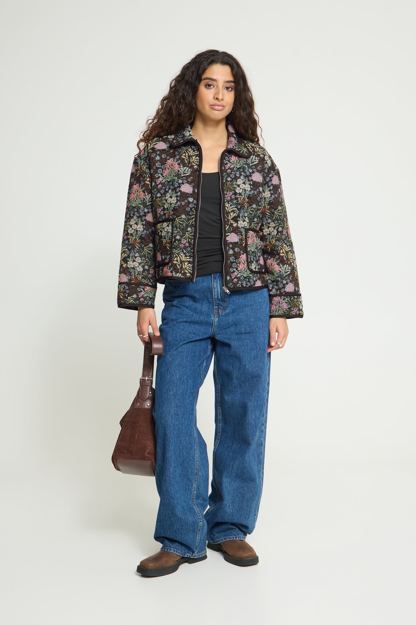 A woman stands against a light gray studio background wearing a cropped floral jacket over a black top, blue wide-leg jeans, brown shoes, and holding a brown shoulder bag.