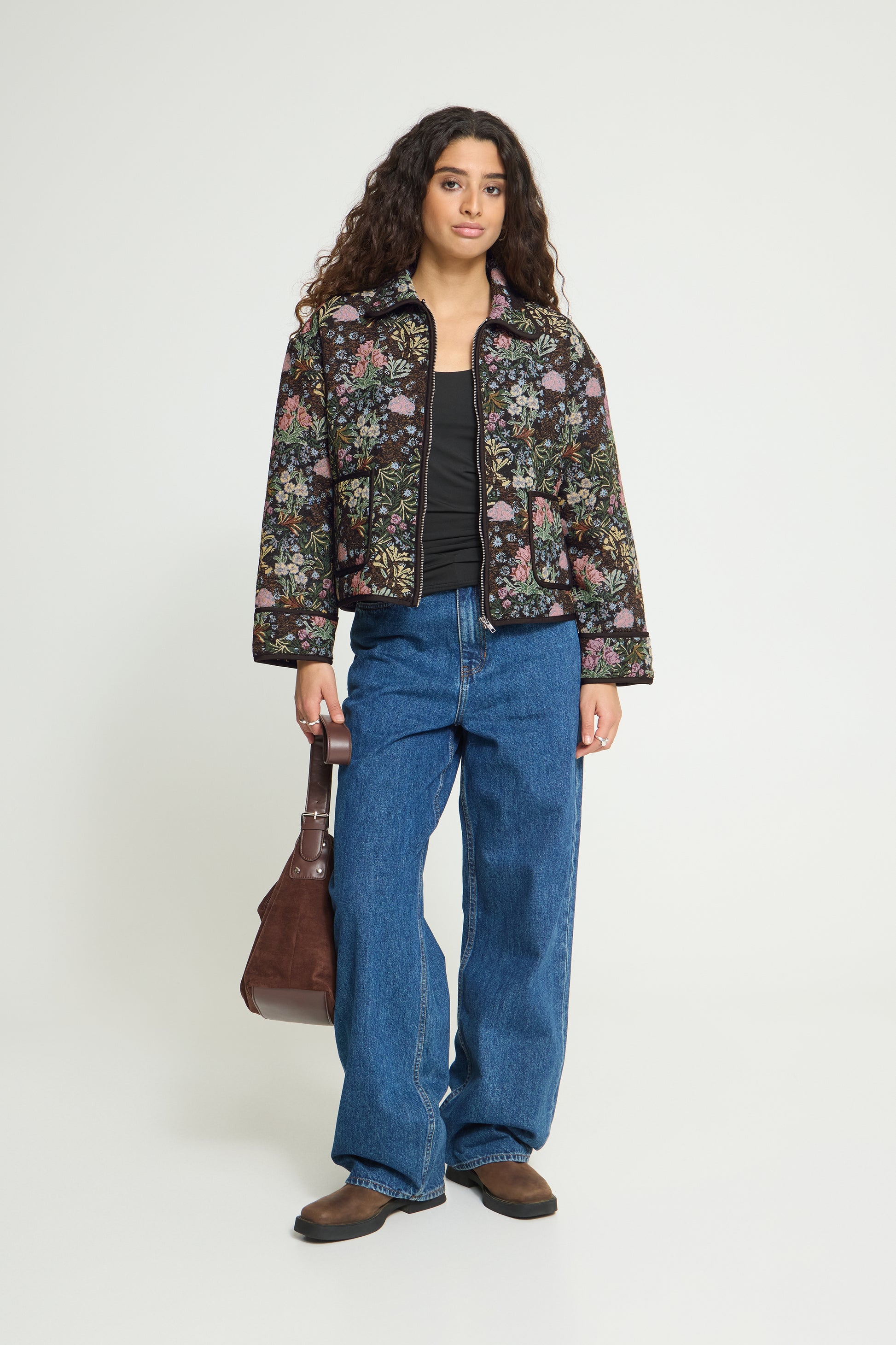 A woman stands against a light gray studio background wearing a cropped floral jacket over a black top, blue wide-leg jeans, brown shoes, and holding a brown shoulder bag.