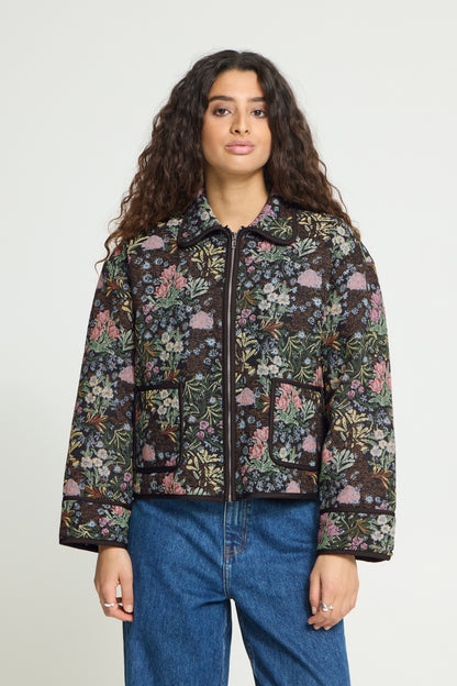 A woman with long curly dark hair models a cropped, dark brown floral quilted jacket with pink and green accents, paired with blue jeans, against a plain light gray background.