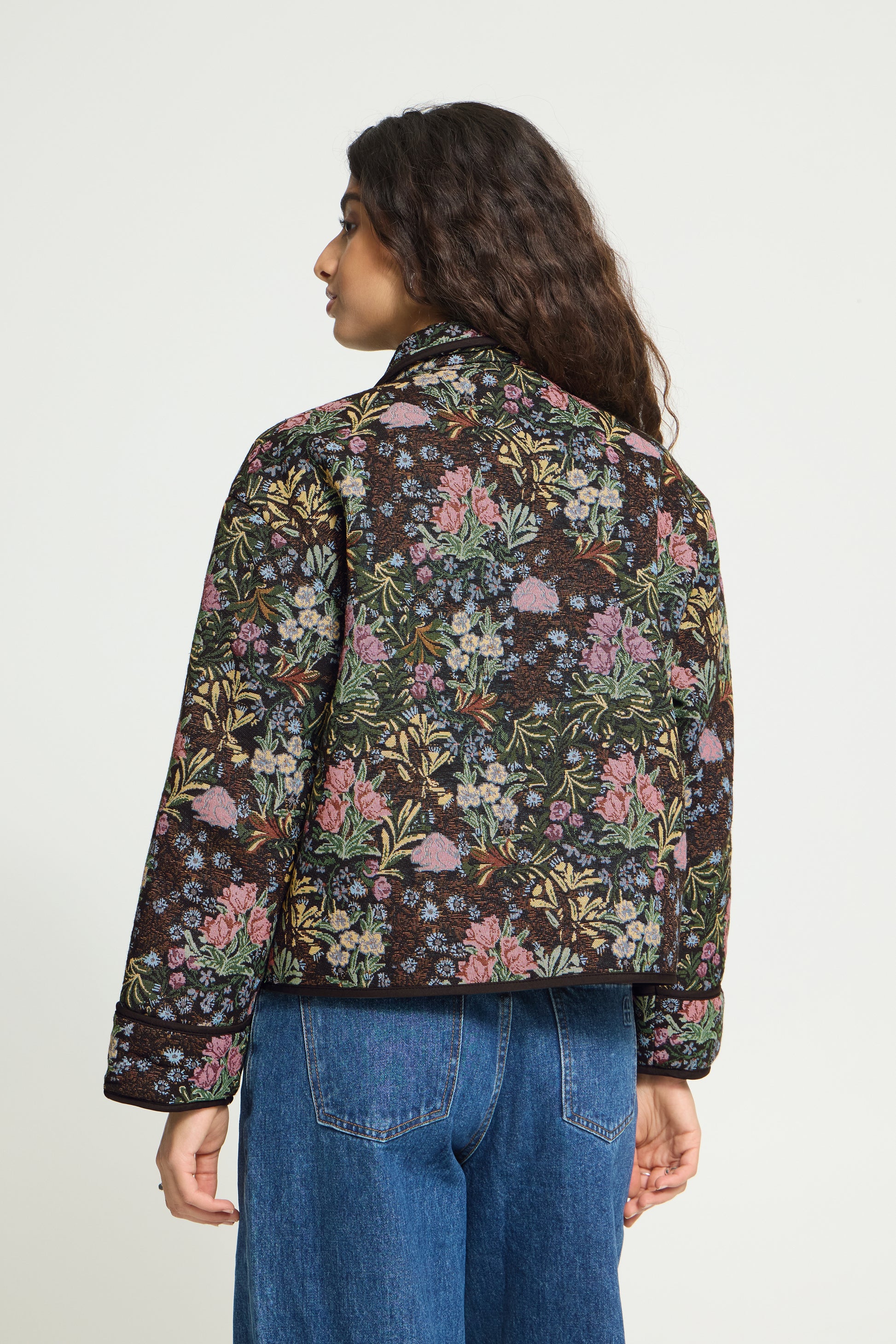 A woman shown from the back wearing a dark brown floral quilted jacket with pink, green, and blue flowers, paired with blue jeans, against a plain white background.