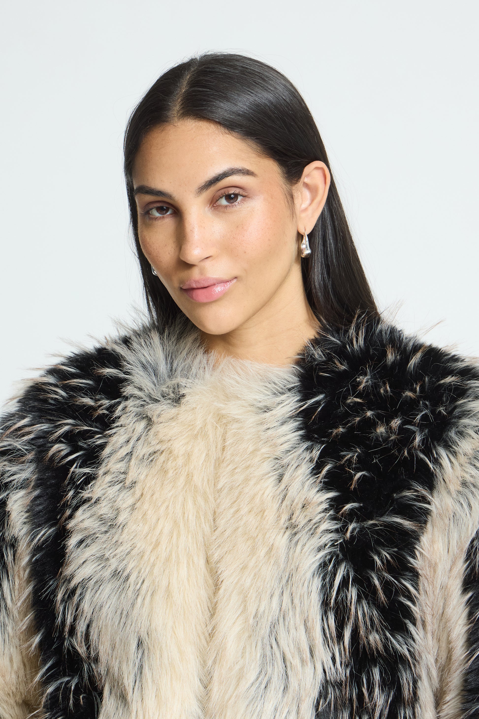 A woman with long dark hair wearing a black and cream faux fur jacket against a light gray studio background.