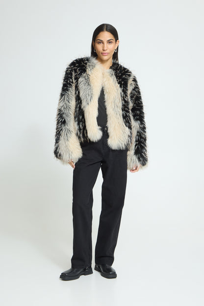 A model stands against a light gray studio background wearing a black and white mottled faux fur jacket over a black top and black trousers.