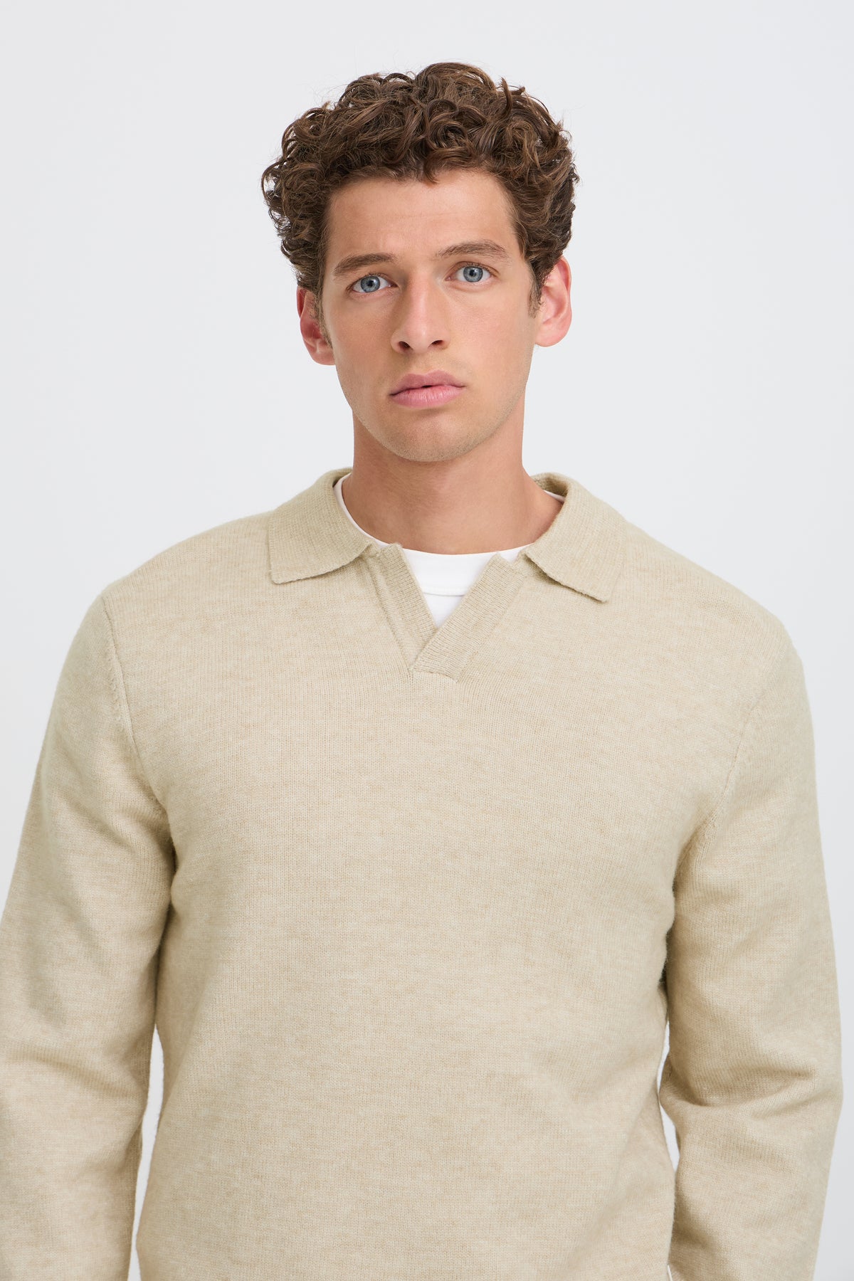 A man wearing a light beige knit polo sweater over a white undershirt, photographed against a clean light gray studio background.