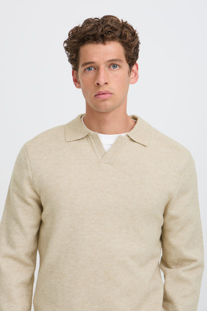 A man wearing a light beige knit polo sweater over a white undershirt, photographed against a clean light gray studio background.
