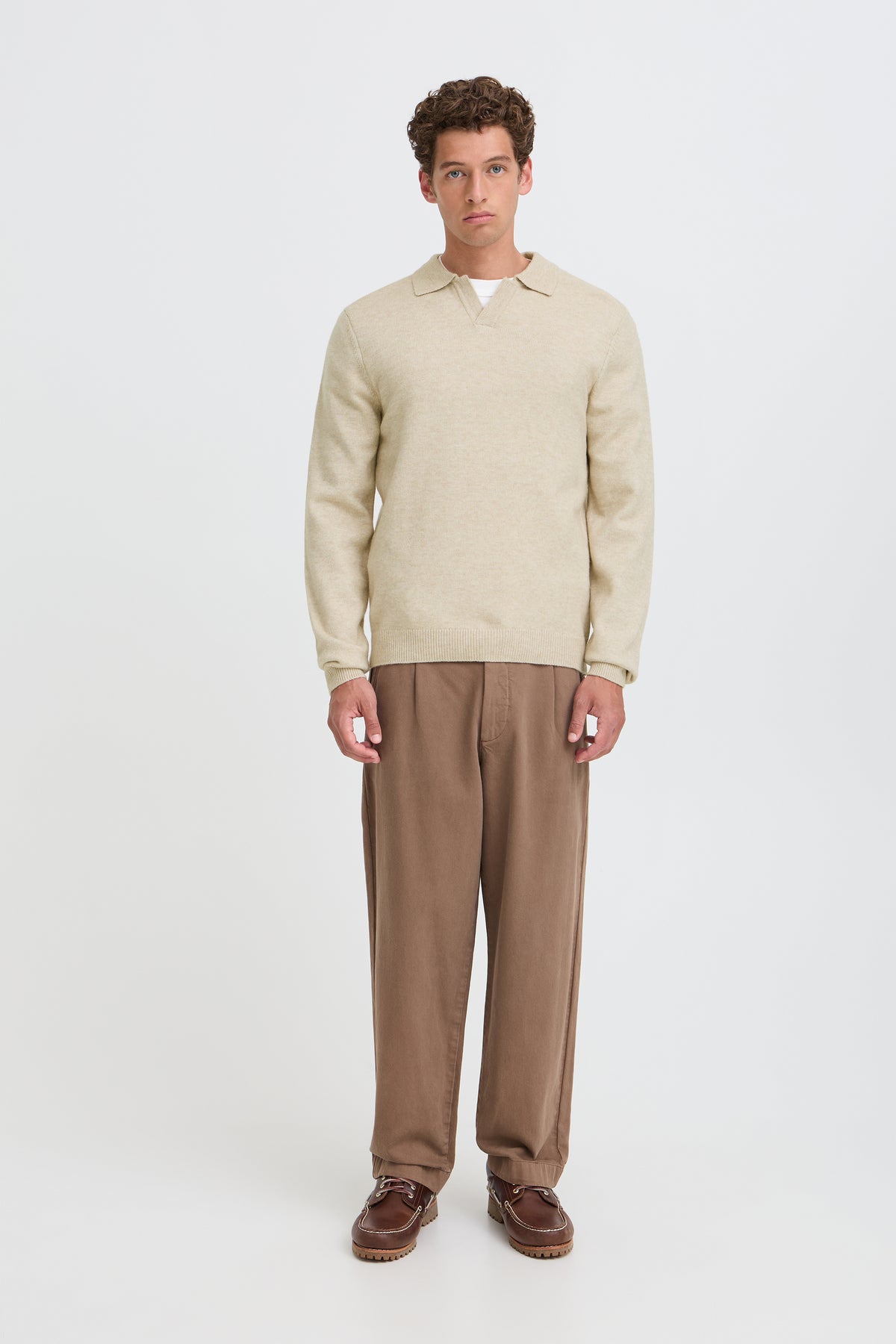 A man stands against a light gray studio background wearing a beige knit polo sweater, brown trousers, and brown shoes.