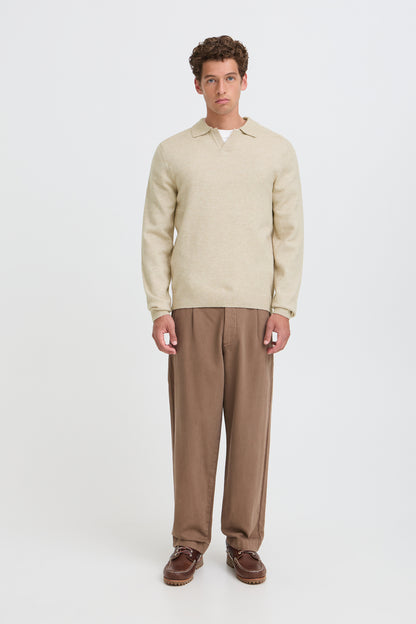 A man stands against a light gray studio background wearing a beige knit polo sweater, brown trousers, and brown shoes.