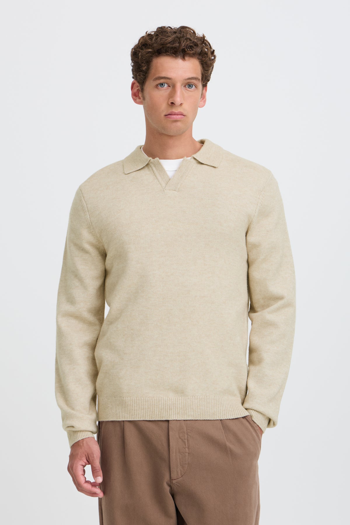 A man wearing a light beige knit polo sweater over a white undershirt and brown trousers, standing against a plain light gray studio background.