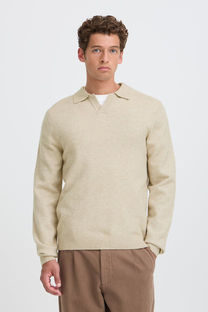 A man wearing a light beige knit polo sweater over a white undershirt and brown trousers, standing against a plain light gray studio background.