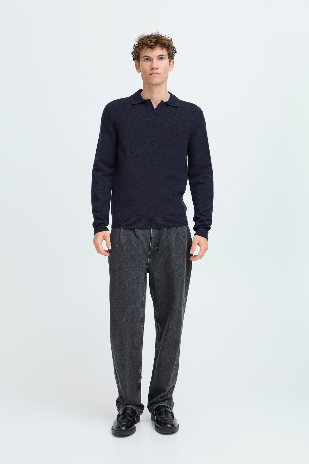 A man modeling a dark navy long-sleeve polo shirt paired with dark gray trousers and black shoes, standing against a plain light gray studio background.