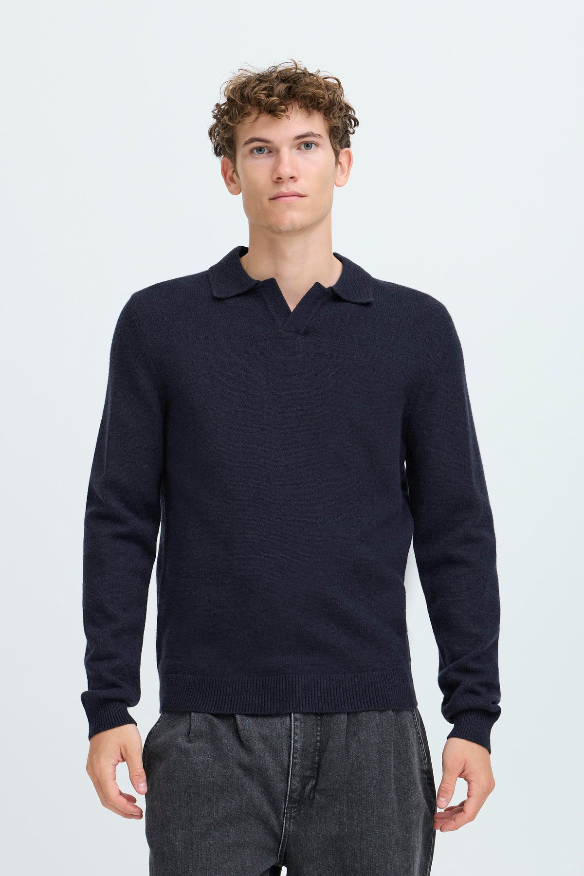A man wearing a dark navy blue long-sleeve knit polo sweater and dark gray jeans, standing against a light gray studio background.