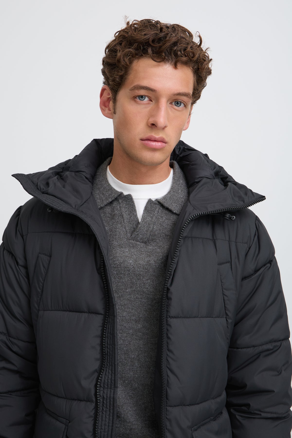 A man wearing a black puffer jacket over a gray sweater and white t-shirt, photographed against a light gray studio background.