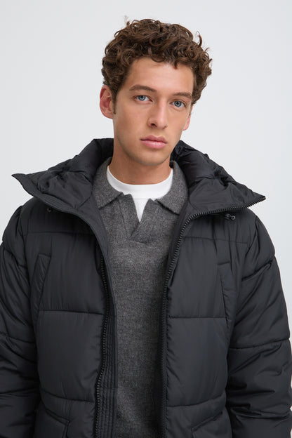 A man wearing a black puffer jacket over a gray sweater and white t-shirt, photographed against a light gray studio background.