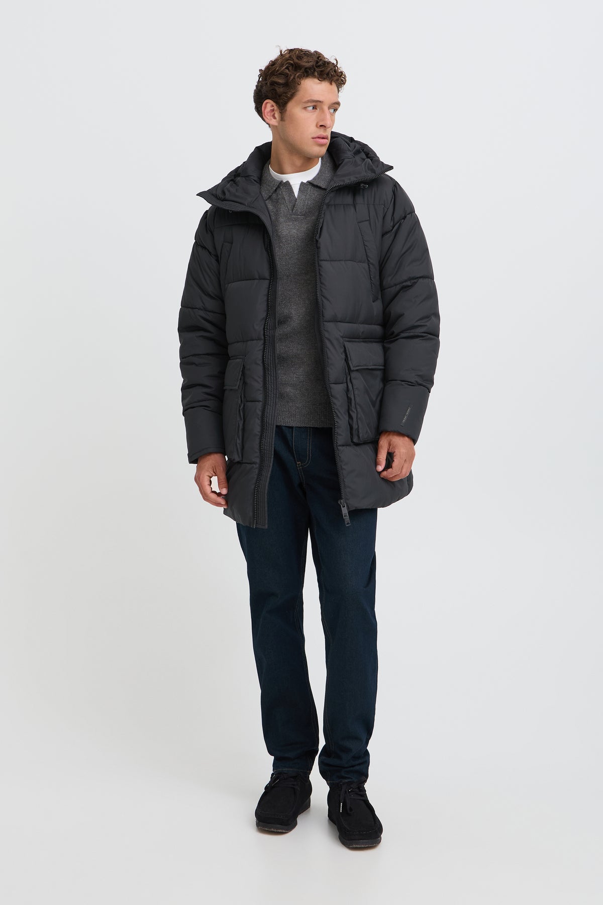 A man modeling a black hooded puffer jacket over a gray sweater, dark blue jeans, and black sneakers, standing against a light gray studio background.