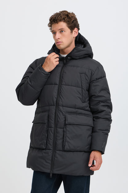 A man wearing a black hooded puffer jacket with front flap pockets, shown against a plain light gray studio background.