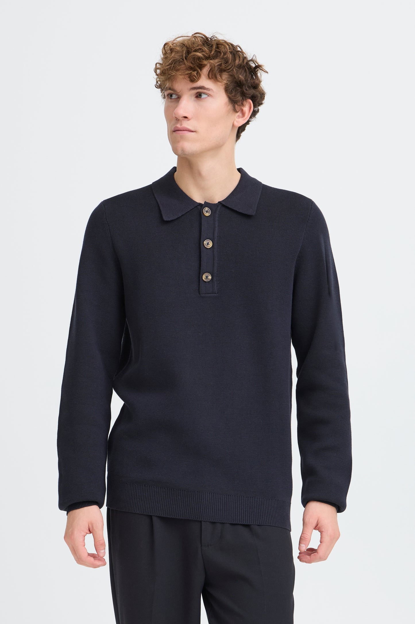 A man wearing a dark navy long-sleeve polo sweater with a three-button placket, paired with dark trousers, photographed against a light gray studio background.