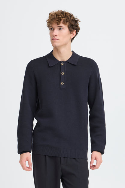 A man wearing a dark navy long-sleeve polo sweater with a three-button placket, paired with dark trousers, photographed against a light gray studio background.