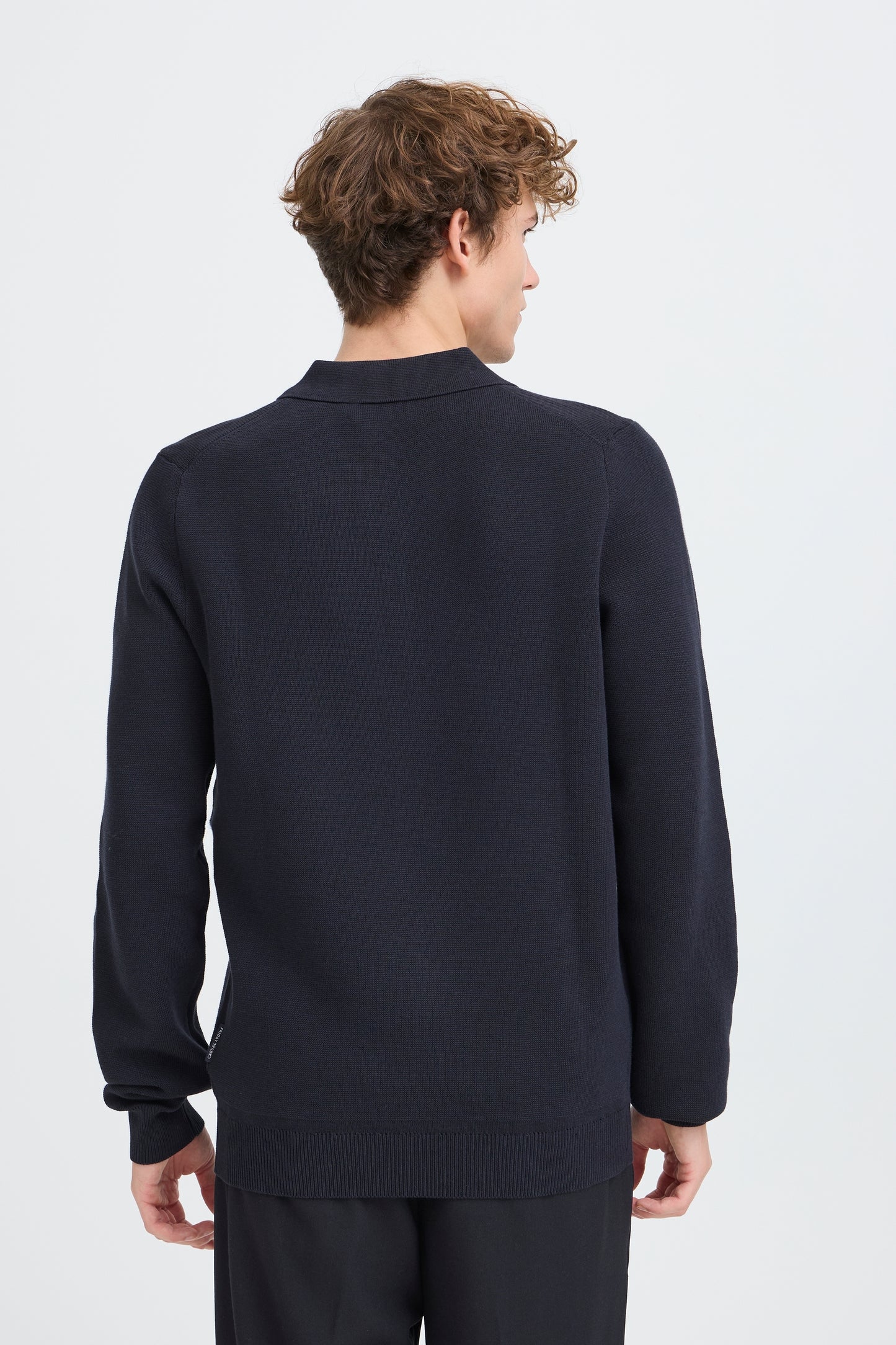 A man viewed from the back wearing a dark navy long-sleeve knit sweater and matching dark trousers against a light gray studio background.