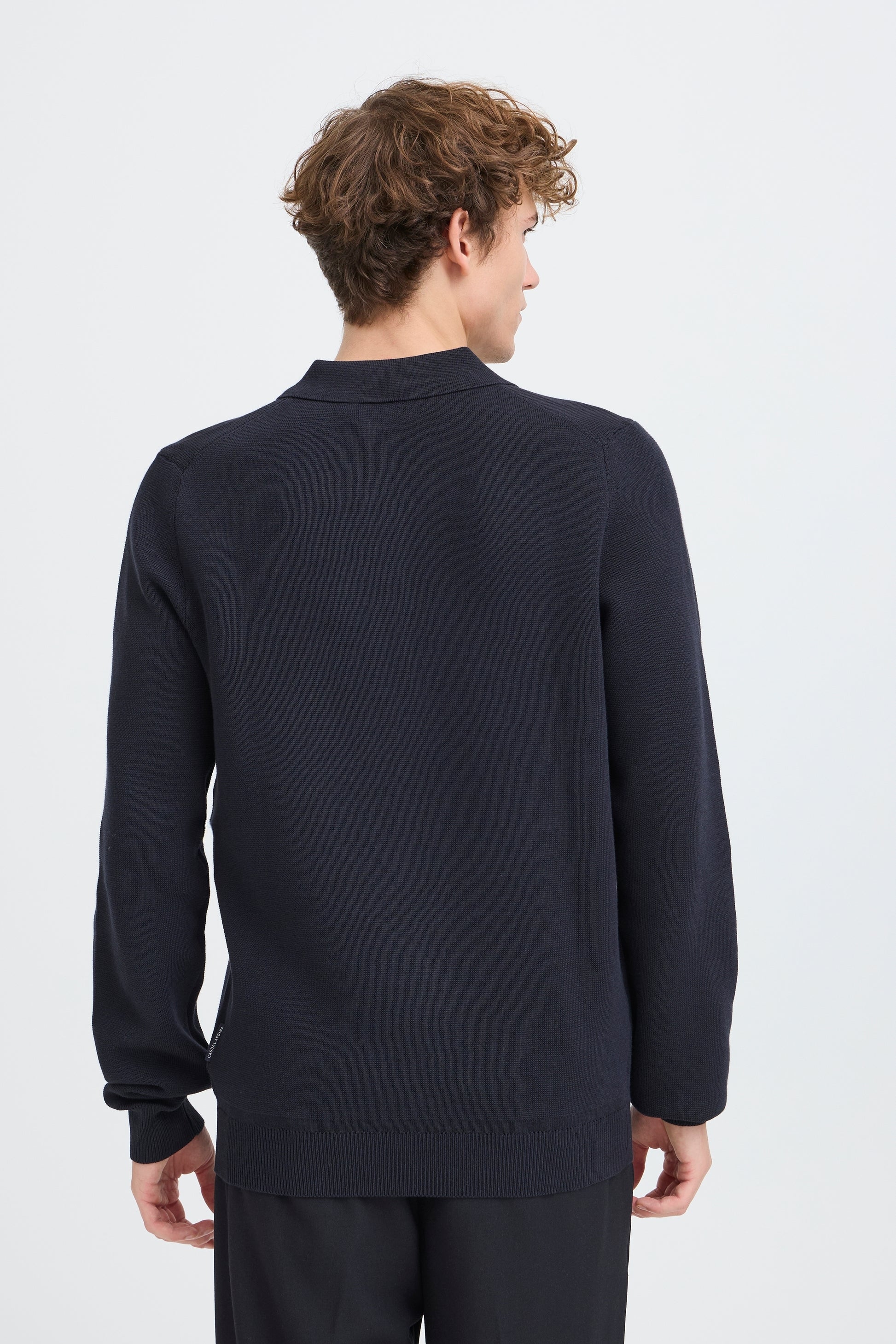 A man viewed from the back wearing a dark navy long-sleeve knit sweater and matching dark trousers against a light gray studio background.
