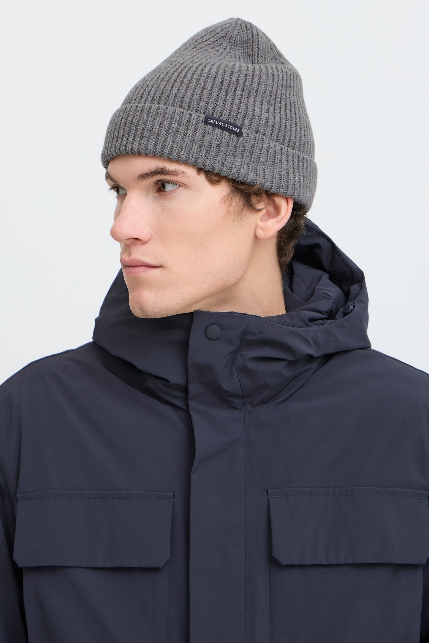 A man wearing a gray ribbed knit beanie and a dark navy hooded jacket, shown in profile against a light gray studio background.