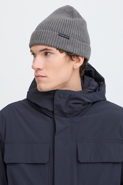 A man wearing a gray ribbed knit beanie and a dark navy hooded jacket, shown in profile against a light gray studio background.