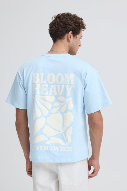 Person wearing a light blue t-shirt with 'BLOOM' text and leaf design on the back, standing against a plain background.