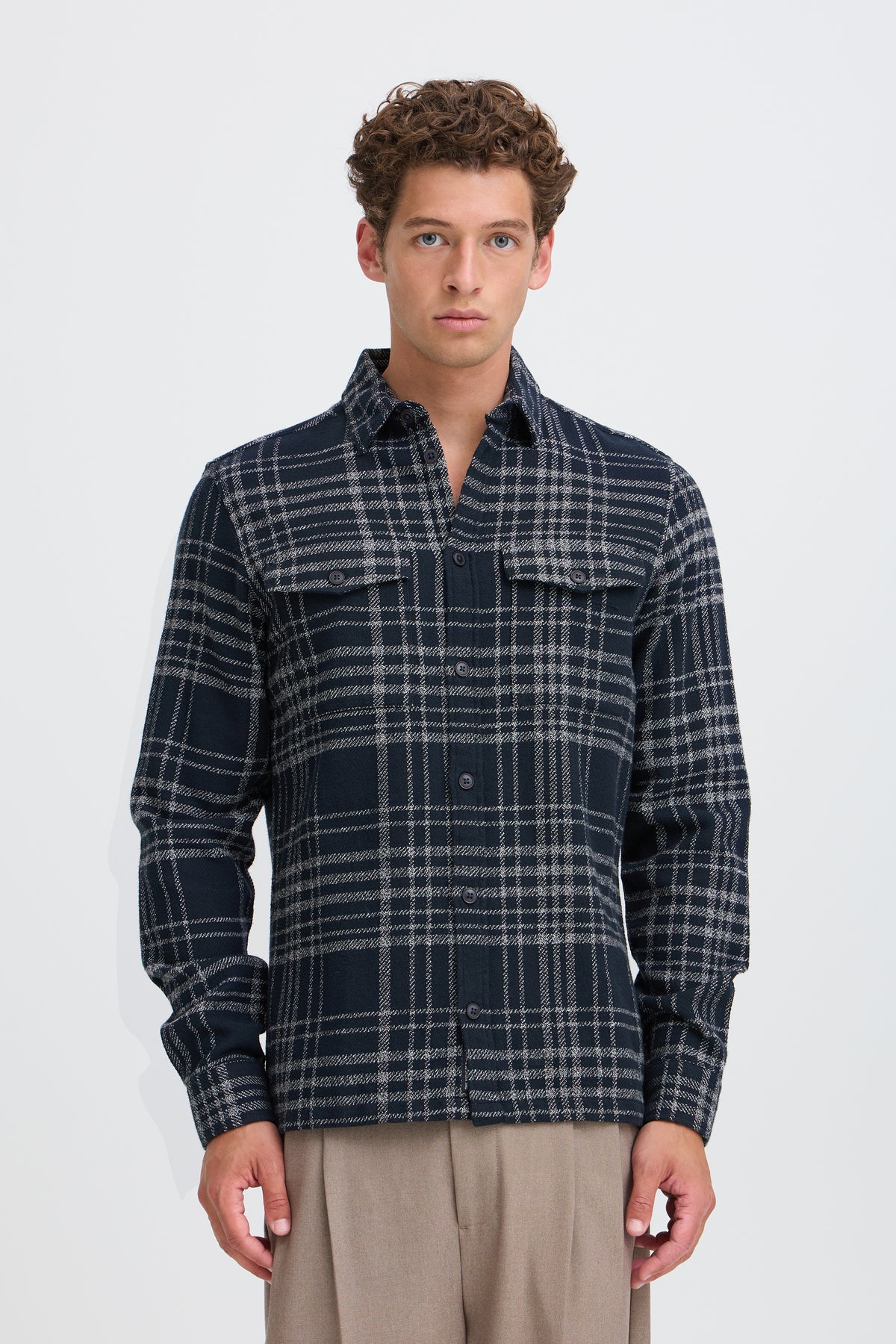 A man wearing a dark navy and gray plaid button-up shirt with two chest pockets, paired with beige trousers, standing against a light gray studio background.