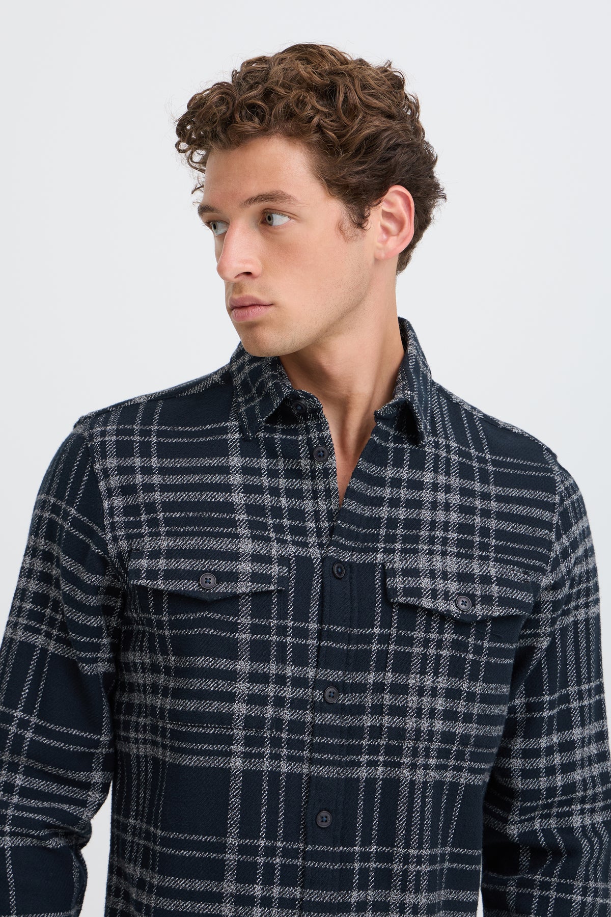 A man wearing a dark navy and gray plaid button-down shirt, shown from the chest up against a light gray studio background.