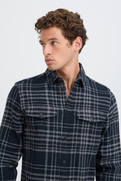 A man wearing a dark navy and gray plaid button-down shirt, shown from the chest up against a light gray studio background.