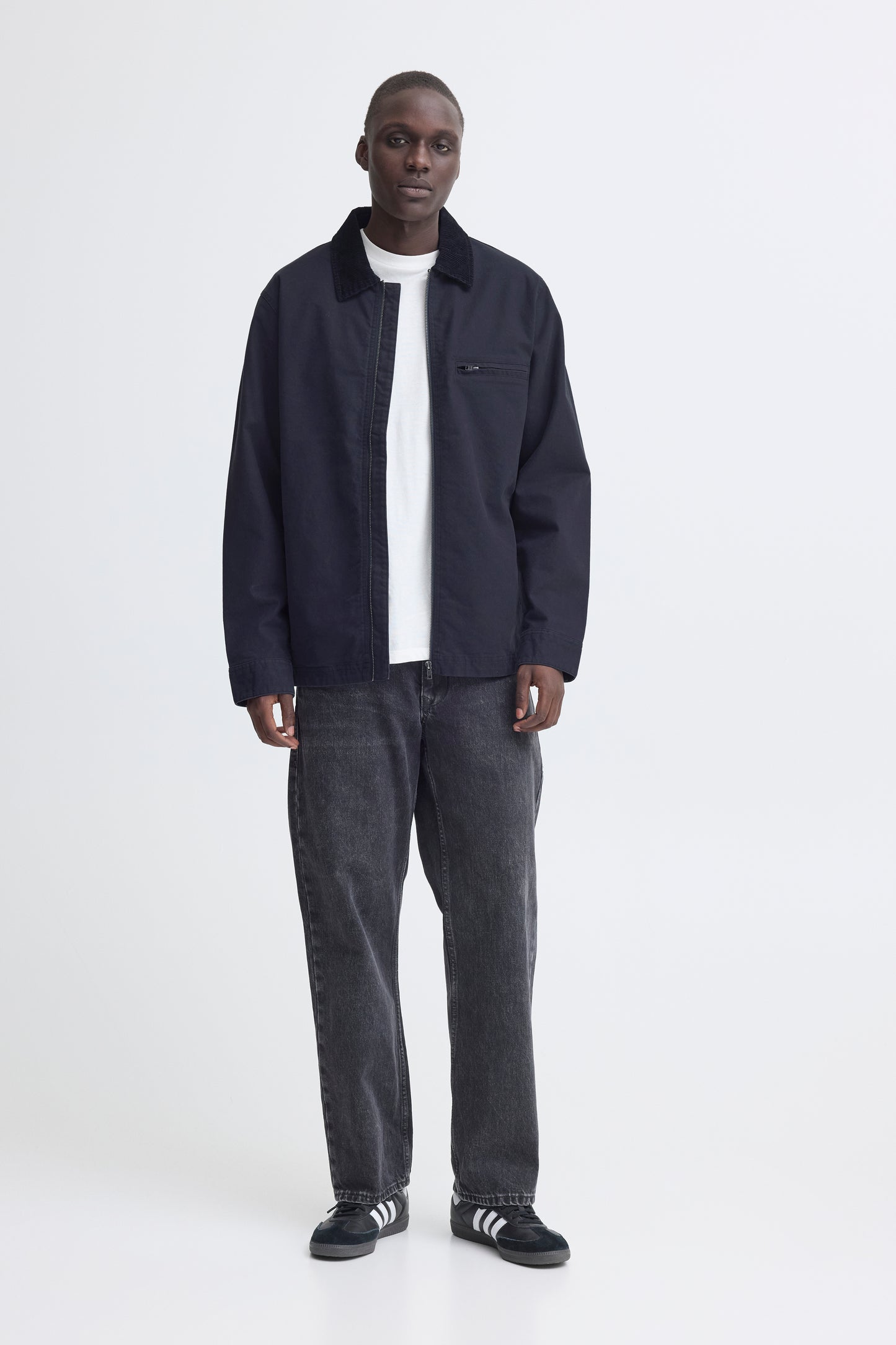 Man wearing a dark jacket, white shirt, gray jeans, and black shoes on a white background