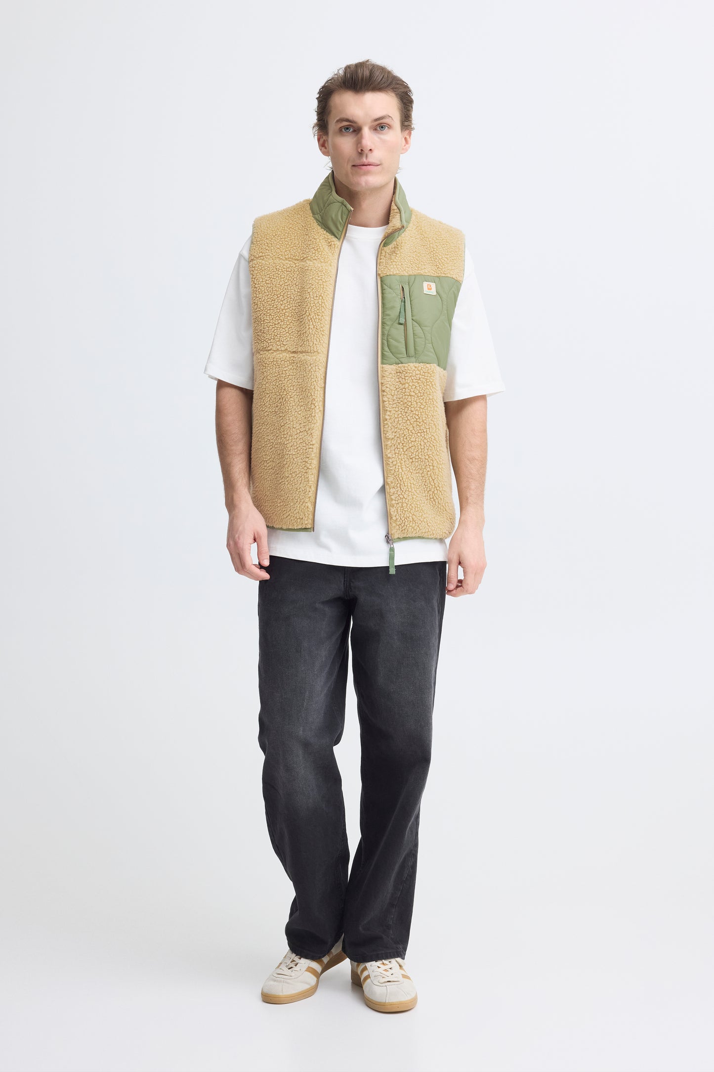 Person wearing a beige and green vest over a white shirt with dark pants on a white background
