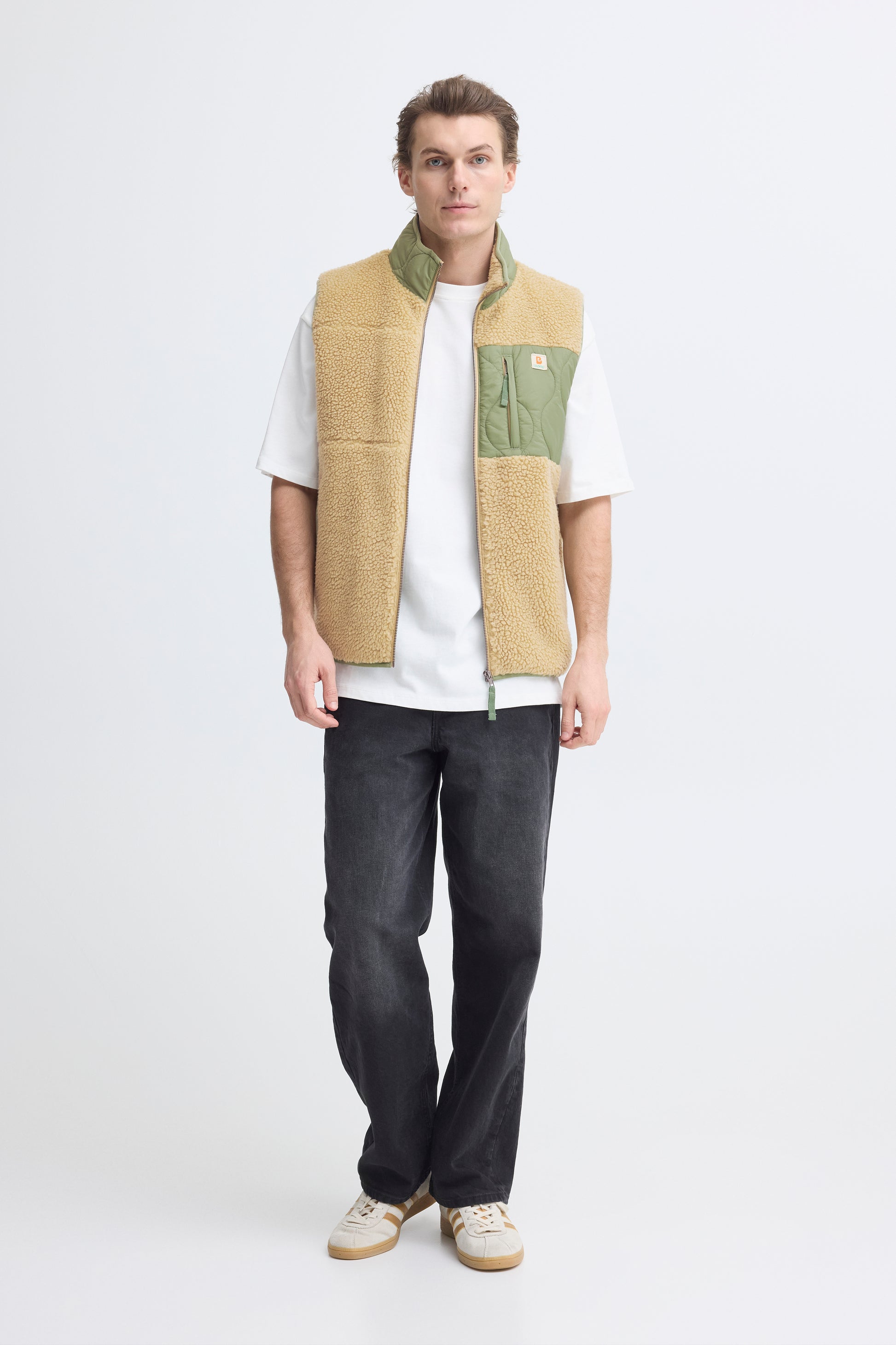 Person wearing a beige and green vest over a white shirt with dark pants on a white background
