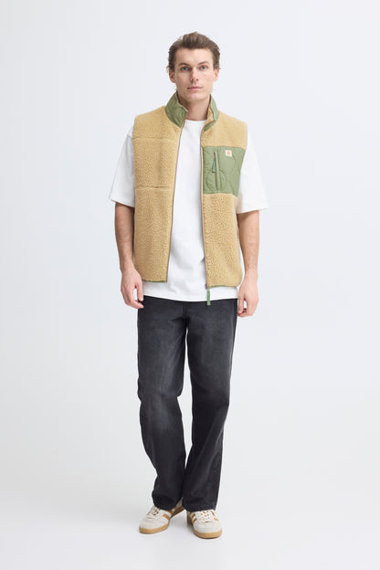 Person wearing a beige and green vest over a white shirt with dark pants on a white background