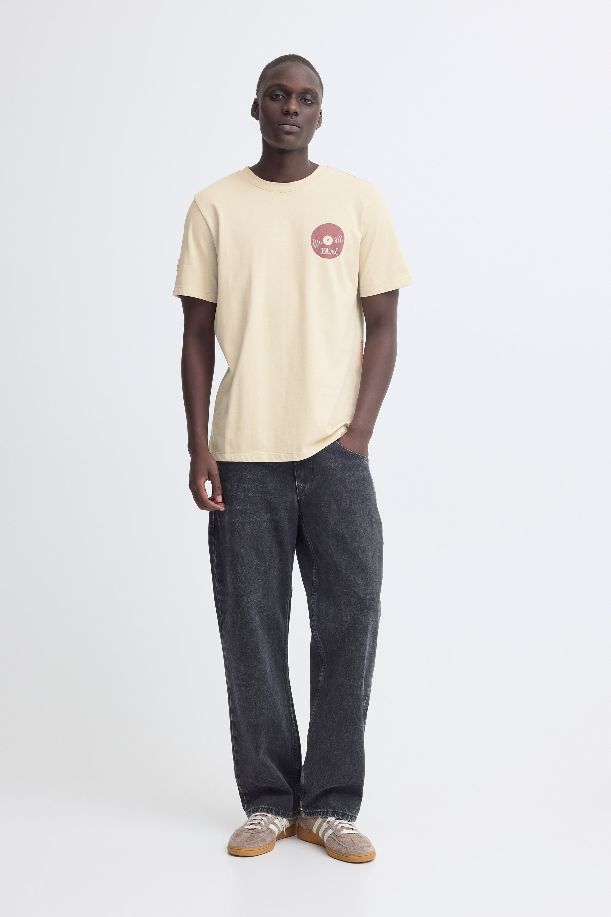 Man wearing a beige t-shirt with a logo and dark jeans on a white background