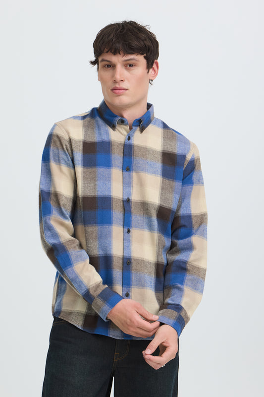 A man wearing a blue, beige, and brown plaid button-down shirt, paired with dark jeans, standing against a light gray studio background.