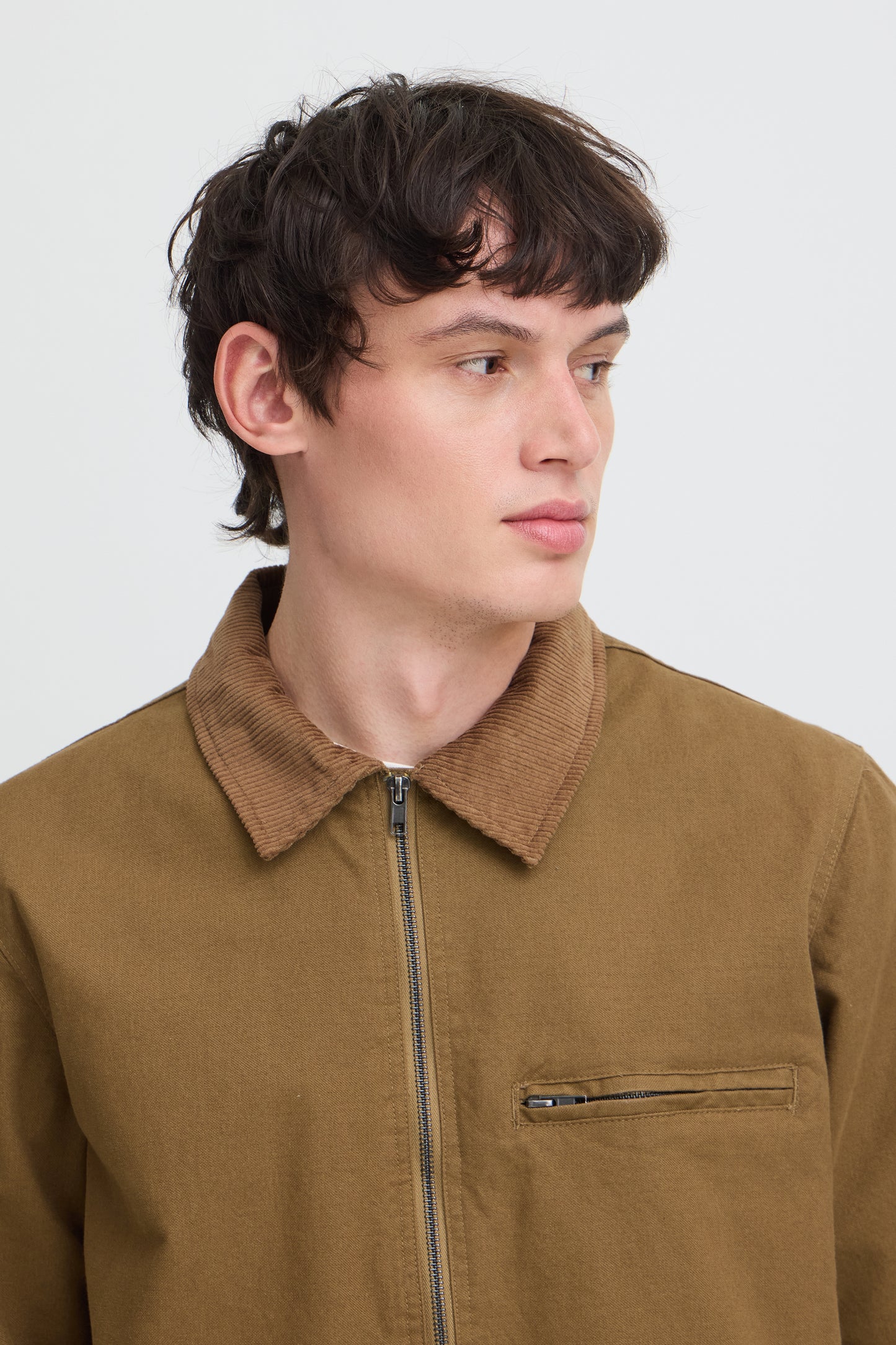 A close-up shot of a man wearing a brown corduroy collar jacket with a front zipper and a zippered chest pocket, set against a plain light gray background.