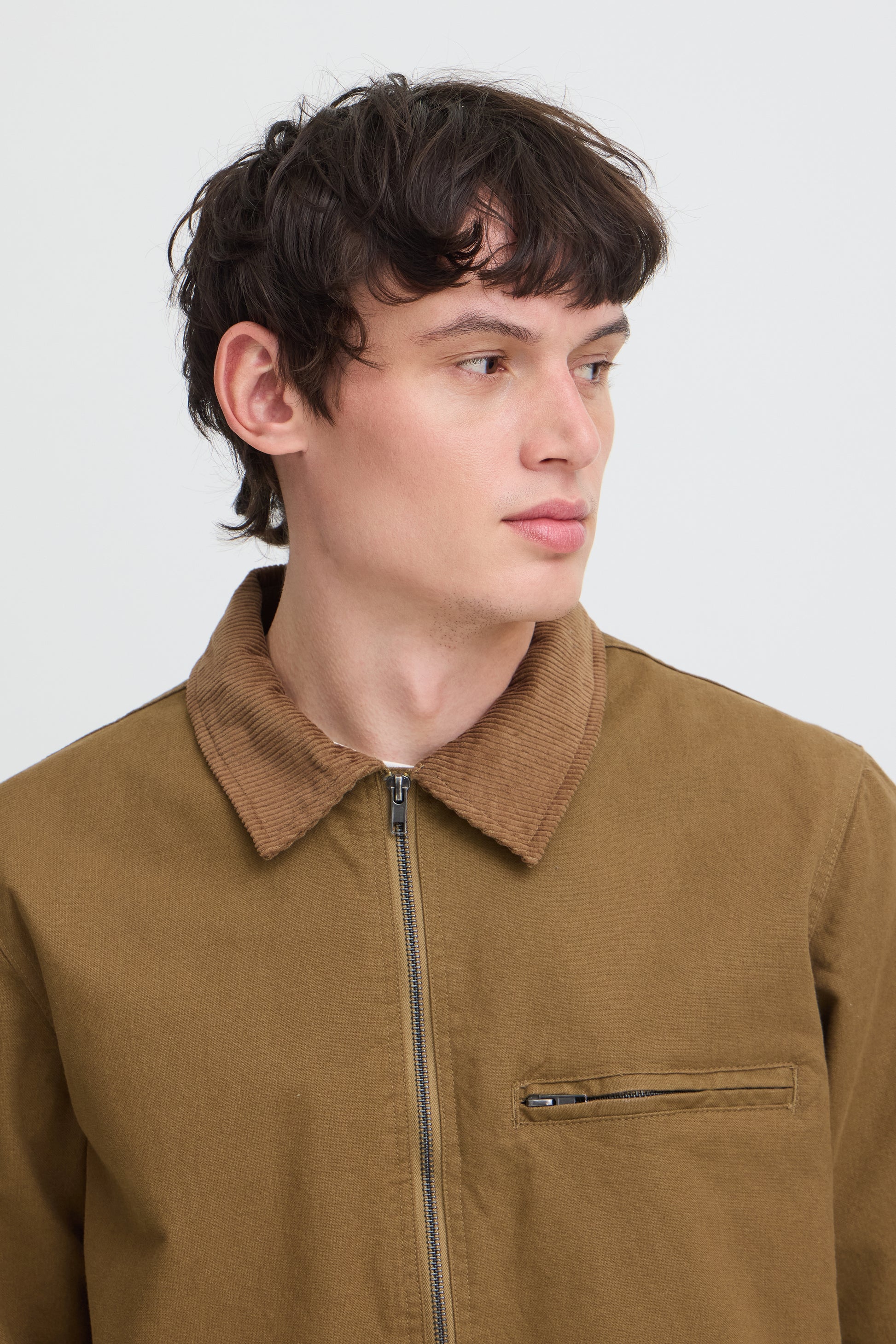 A close-up shot of a man wearing a brown corduroy collar jacket with a front zipper and a zippered chest pocket, set against a plain light gray background.