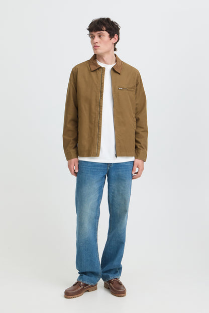A man modeling a brown overshirt jacket over a white t-shirt, paired with blue jeans and brown leather shoes, standing against a light gray studio background.